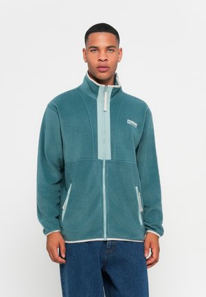 Fleecejacka i teal med hög krage, framdragkedja och kontrasterande ljusgröna paneler. Har sidofickor och broderade detaljer.