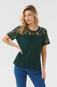 Top in pizzo verde scuro con maniche corte, scollatura ondulata e motivo floreale intricato; abbinato a jeans di denim.