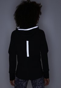 Un hoodie noir en tissu texturé, avec une bande réfléchissante dans le dos et des finitions blanches contrastantes sur la capuche.