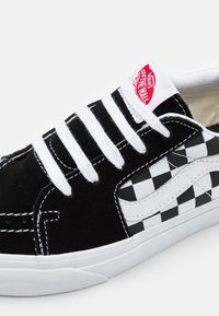 Black and white checkerboard canvas sneaker with a suede toe, white laces, white rubber sole, and a red Vans logo on the tongue.