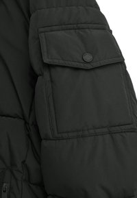 Mörkgrön pufferjacka med en ärm som har en sydd ficka med en rund svart knapp märkt "Polo Club". Slät yta och quiltad design.