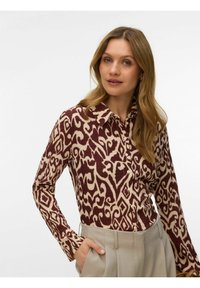 Femme aux longs cheveux blonds portant un pantalon beige taille haute et une chemise à manches longues boutonnée avec un motif marron et crème, main dans la poche.