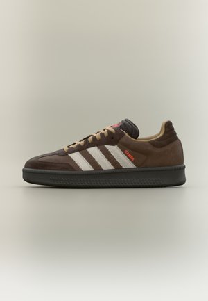 Zapatilla Adidas Samba marrón con rayas y cordones beige, suela negra y texto rojo "SAMBA" en el lateral, mostrada de perfil sobre un fondo neutro.