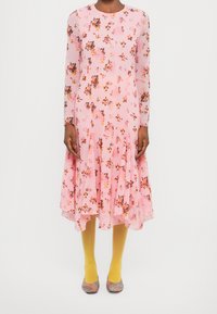 Femme portant une robe fleurie rose à manches longues, des collants jaunes et des chaussures multicolores, debout devant un fond blanc uni.