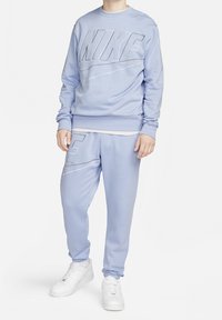 Světle modrá mikina Nike a odpovídající tepláky, oboje vyrobené z jemného materiálu, s velkými logy a volným střihem. Bílá obuv dotváří vzhled.