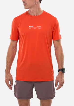 Homme portant un t-shirt à manches courtes orange vif avec un texte blanc et un short gris, debout les bras détendus le long du corps.