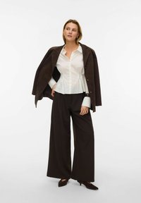 Weißes Hemd mit Knöpfen und Schößchenbund, braune Wildlederjacke lässig über die Schultern gelegt, weite dunkle Hosen und dunkle spitze Schuhe.