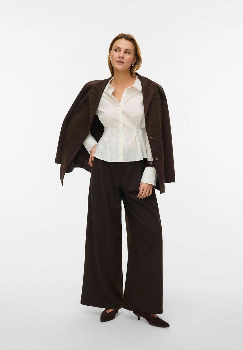 Weißes Hemd mit Knöpfen und Schößchenbund, braune Wildlederjacke lässig über die Schultern gelegt, weite dunkle Hosen und dunkle spitze Schuhe.