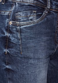 Jeans blu scuro con una texture liscia, caratterizzati da cuciture gialle a contrasto e una piccola tasca anteriore con un dettaglio a bottone argentato.