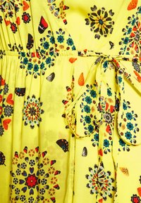 Geel fabric met gekleurde vlinder- en mandala-patronen, met een gegatherde taille en een strikdetail van stof.