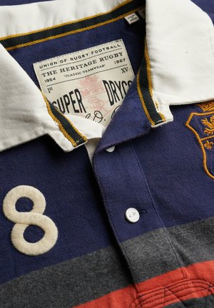 Rugbyshirt mit weißem Kragen, blauen, grauen und roten horizontalen Streifen, eingestickter Nummer 8 und einem gelben gestickten Wappen auf der Brust.