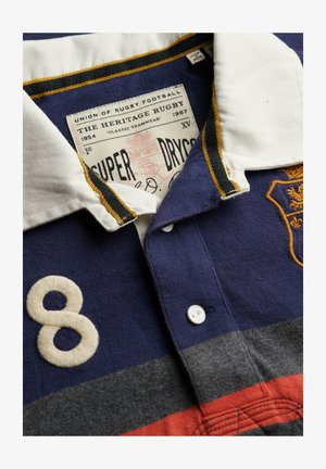 Rugbyshirt mit weißem Kragen, blauen, grauen und roten horizontalen Streifen, eingestickter Nummer 8 und einem gelben gestickten Wappen auf der Brust.