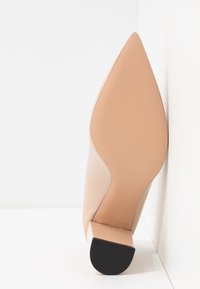 Escarpin de style stiletto en cuir verni nude avec un bout pointu. La semelle est noire et la chaussure présente une texture lisse et brillante.