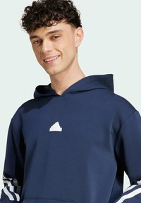 Námořnická modrá mikina s kapucí a bílým logem Adidas na hrudi. Má kapuci a bílé pruhy na rukávech. Měkká textura, pohodlný design.