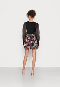 Top de malha preto com mangas bufantes transparentes, combinado com uma saia mini floral em camadas em cores vibrantes sobre um fundo escuro. Sapatos de salto nude completam o visual.