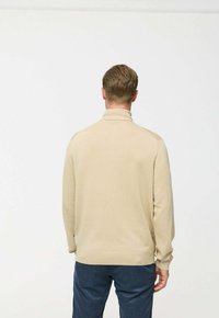 Beige kötött pulóver magas gallérral és bordázott mandzsettákkal, laza szabással. Sötétkék nadrág felett viselik, hátulról nézve.