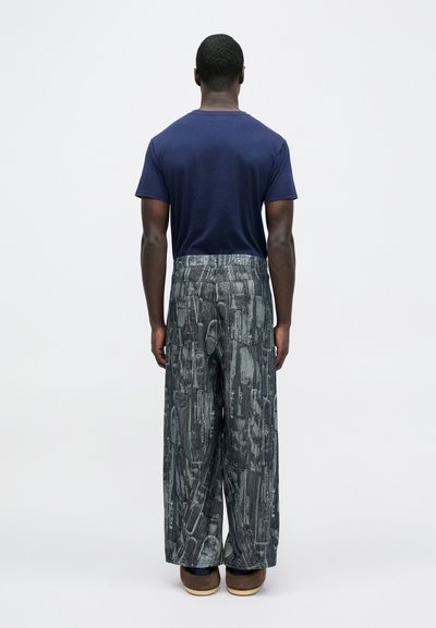 Μπλε ναυτικό κοντομάνικο t-shirt σε συνδυασμό με φαρδιά παντελόνια με σχέδια σε γκρι αποχρώσεις, που διαθέτουν αφηρημένα μοτίβα και υφή. Καφέ παπούτσια.