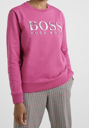 Osoba nosząca jaskraworóżową, długorękawową bluzę z nadrukiem „BOSS HUGO BOSS” w srebrnych literach oraz wzorzyste spodnie w kratę, dłonie zrelaksowane.