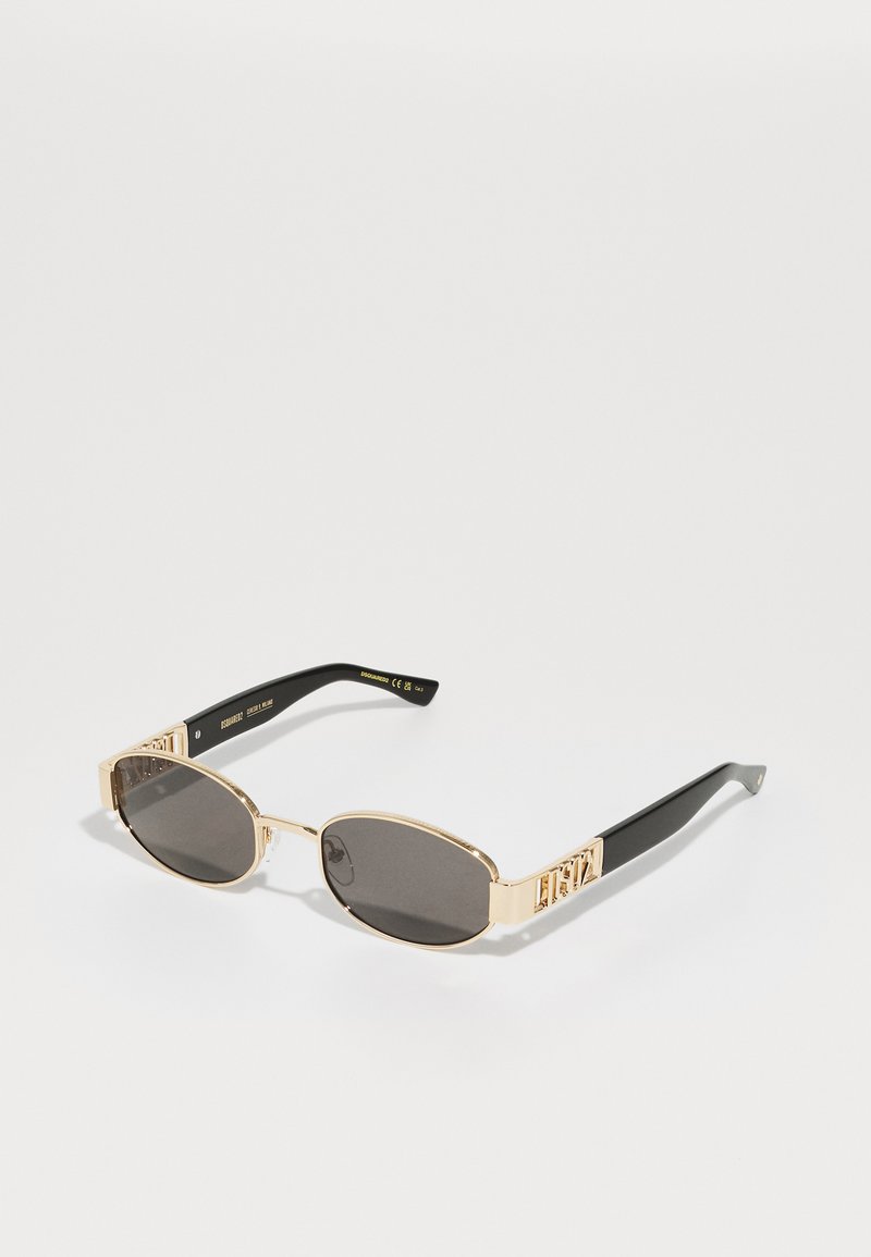 Occhiali da sole con montatura dorata, lenti scure, forma ovale e aste nere con un distintivo dettaglio del logo dorato.