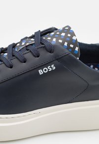 Baskets bleu marine avec lacets texturés, logo blanc "BOSS" sur le côté, languette à motif géométrique bleu, blanc et noir, et semelle blanche épaisse.