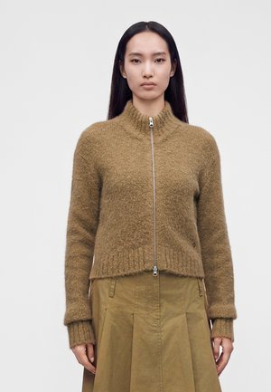 Donna con capelli lunghi e scuri che indossa un maglione marrone peloso con zip e una gonna verde oliva, in piedi davanti a uno sfondo semplice.