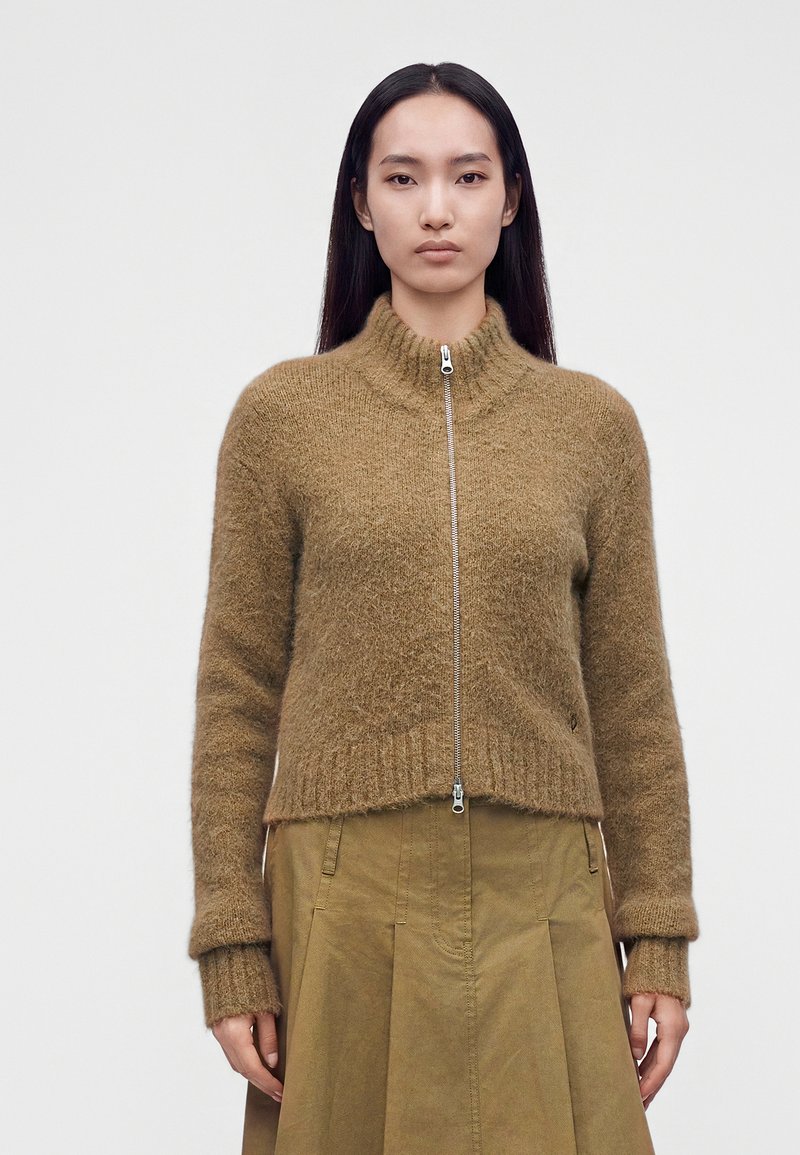 Femme aux longs cheveux foncés portant un pull marron en tissu pelucheux avec fermeture éclair et une jupe vert olive, debout devant un fond uni.