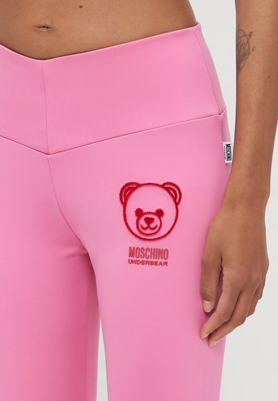 Ružové legíny vyrobené z hladkej tkaniny, s červeným vyšitým logom medvedíka a textom "Moschino Underbear" na pravej hornej stehne.