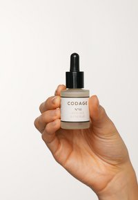 CODAGE N°10 Anti-Aging-Serum in einer matten Glastropfflasche mit schwarzem Deckel; weißes Etikett mit schwarzem Text, in einer Hand gehalten.