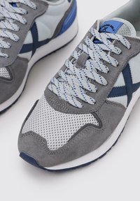 Zapatos deportivos en gris y azul claro con materiales de malla y gamuza, presentan cordones con patrones, una suela blanca y un acento azul en el talón.