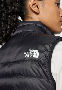 Crna puffer prsluk s visokim ovratnikom, s bijelim logotipom "The North Face" na gornjem dijelu leđa. Glatka, reflektirajuća tekstura.