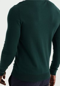 Man som bär en mörkgrön långärmad tröja och mörka byxor, visad bakifrån mot en enfärgad bakgrund.