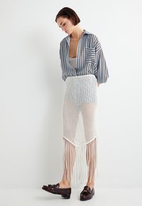 Chemise oversize rayée bleue et blanche associée à une jupe en maille blanche avec de longues franges. Modèle portant des mocassins noirs.
