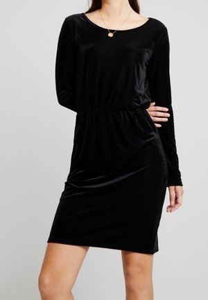 Frau trägt ein knielanges schwarzes Samtkleid mit langen Ärmeln, rundem Ausschnitt und einer goldenen Anhängerkette.