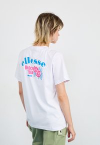T-shirt en coton blanc avec un imprimé graphique color�é au dos avec "ellesse", "Dolce Vita" et des accents floraux. Manches courtes, coupe décontractée.