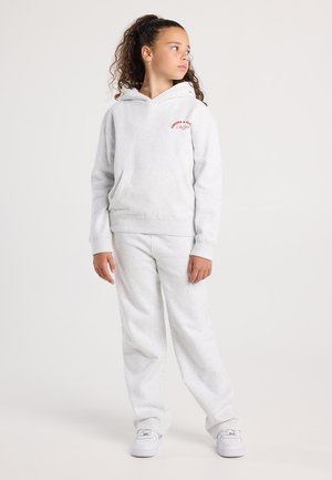 Mädchen mit lockigem Haar in einem weißen Hoodie und Jogginghose, stehend und zur Seite schauend vor einem schlichten hellen Hintergrund.