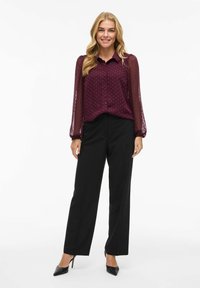 Chemise bordeaux à boutons avec des manches longues transparentes et un motif à pois, associée à un pantalon noir coupe droite et des talons noirs.