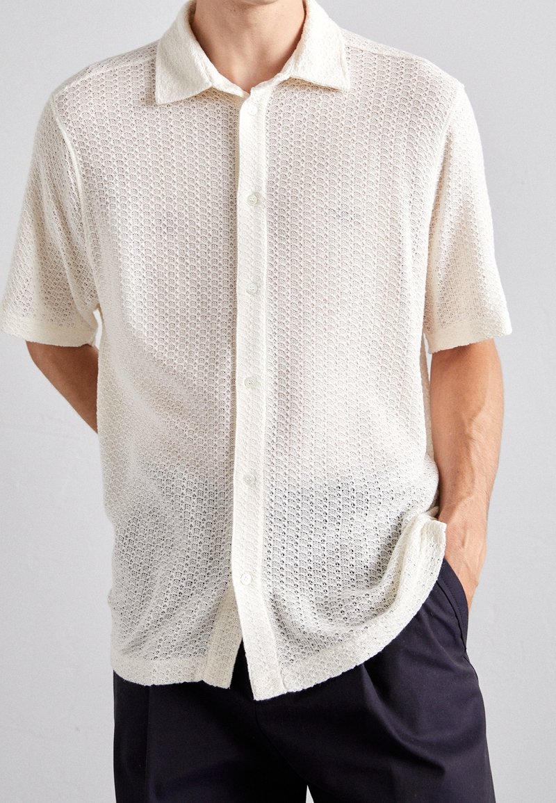 Camisa blanca de manga corta con un patrón texturizado de panal, con cierre de botones en el frente y un cuello suave, llevada sobre pantalones oscuros.
