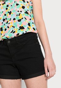 Calções de ganga pretos com a barra dobrada, combinados com uma blusa de estampa floral multicolorida com mangas bufantes. Textura de tecido suave.