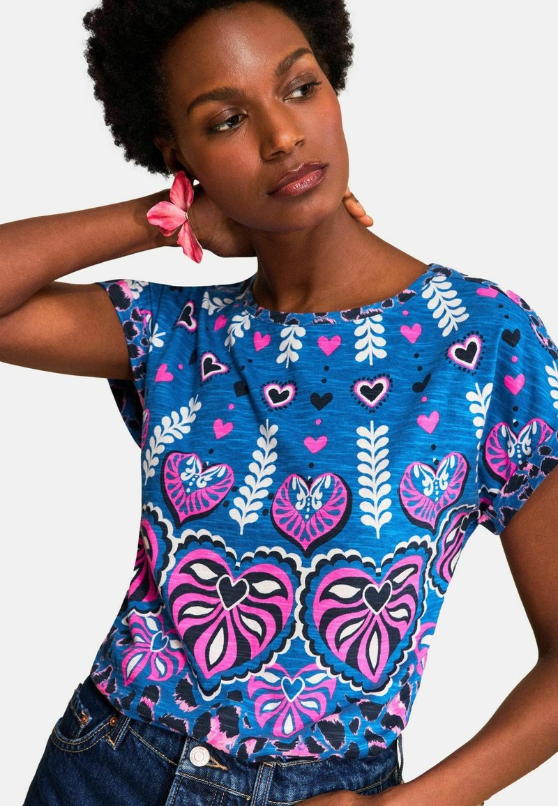 Lühikeste lokkis juustega naine, kes kannab sinist särki roosade ja valgete südame- ning lehemustritega, poseerides käsi kaela taga ja kandmas roosat lillekäevõru.