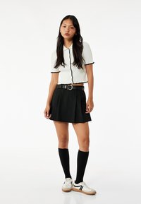 Biały prążkowany crop top z czarnymi akcentami, plisowana czarna spódnica, czarne skarpetki do kolan oraz białe trampki z czarnymi paskami.