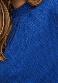 Blue blouse featuring a textured chevron pattern with gathered neckline and flowing fabric. Material appears lightweight and breathable.