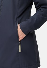 Veste imperméable bleu marine avec une poche zippée. Matériau lisse, design ajusté et accent subtil sur le tirette de la fermeture éclair.