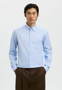 Jonge man in een lichtblauw overhemd met knopen en een bruine geplooide broek, staande met zijn handen in zijn zakken tegen een plain witte achtergrond.