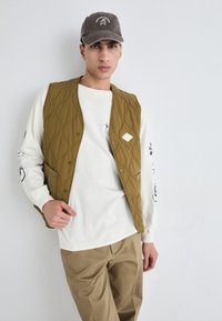 Quilted brown vest with a diamond patch, worn over a white long-sleeve shirt featuring blue accents. Model styled with beige pants and a gray cap.