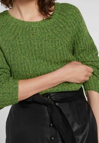 Frau trägt einen grünen strukturierten Strickpullover mit hochgeschobenen Ärmeln und einen schwarzen Lederrock mit gebundenem Gürtel und Knopfdetails.