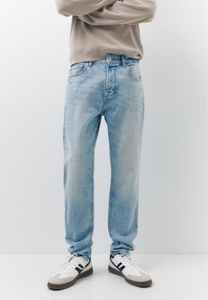 Persoon staand met gekruiste armen, gekleed in lichtblauwe spijkerbroek en witte sneakers met zwarte details, tegen een effen witte achtergrond.