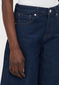 Jeans in denim blu scuro con cinque tasche, cuciture gialle a contrasto, una texture liscia e un taglio dritto.