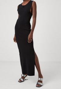 Robe maxi noire sans manches avec une fente latérale, confectionnée dans un tissu lisse. Associée à des sandales à talons argentés brillants avec des accents de boucles.