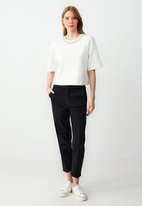 Witte cropped sweatshirt met een versierde halslijn, gecombineerd met zwarte tapered broek en witte sneakers, tegen een neutrale achtergrond.