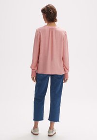 Blouse rose à manches longues avec un dos froncé, associée à un jean bleu court. La blouse présente des poignets texturés froncés et une coupe décontractée.